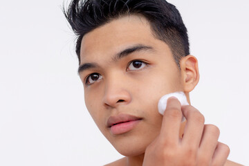 A young asian man using a cotton ball dipped in astringent or skin toner to cleanse his face. Using a dabbing motion. Skin care and hygiene concepts. Isolated on a white backdrop.