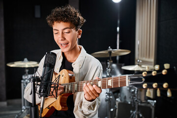 talented cute teenage boy in casual attire playing guitar and singing song into microphone in studio