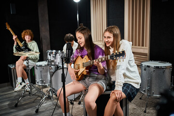 focus on teenage girls with guitar singing with blurred friends with drums and guitar on backdrop