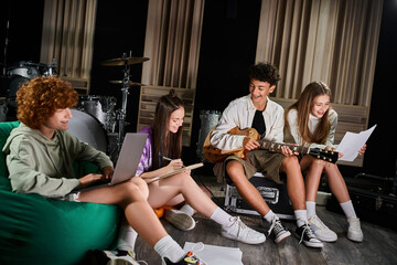 Fototapeta na wymiar four joyous teenage band members in casual attires sitting with guitar and laptop writing lyrics
