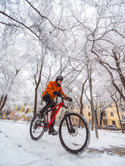 A guy in an orange jacket rides a bicycle through a winter city among snow covered trees. Active lifestyle in winter