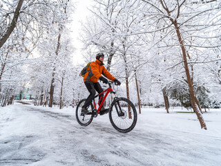 A man in an orange jacket rides a bicycle in a winter park among snow covered trees. Active lifestyle in winter in the city