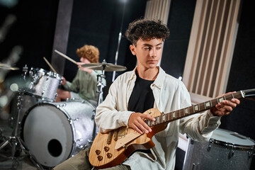 concentrated adorable teenage boy in casual attire playing guitar next to his blurred drummer