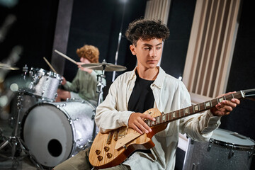 Naklejka premium concentrated adorable teenage boy in casual attire playing guitar next to his blurred drummer