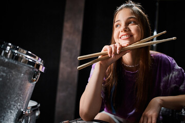 jolly brunette teenage girl in everyday vivid attire posing with drumsticks and looking away