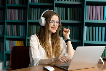 Smiling young woman with headphones using laptop in a library with colorful bookshelves in the background.