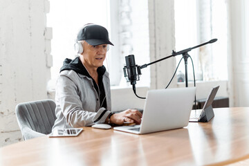 Focused podcaster recording in a studio with a microphone and laptop, wearing headphones and a cap.