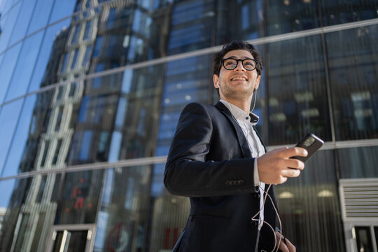 Happy businessman with earphones using smartphone in front of a glass building, enjoying a break.