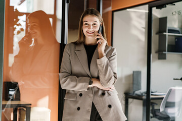 Confident businesswoman in a stylish blazer talking on the phone with office interior reflection.