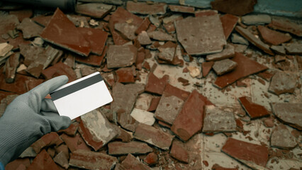woman's hand adorned in construction gloves, holding a bank card, set against a backdrop of shattered tiles on the floor. Symbolizing the initiation of renovation and the considerable expenses