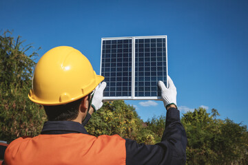 Young male engineer in uniform holding a photovoltaic solar panel on blue sky background.