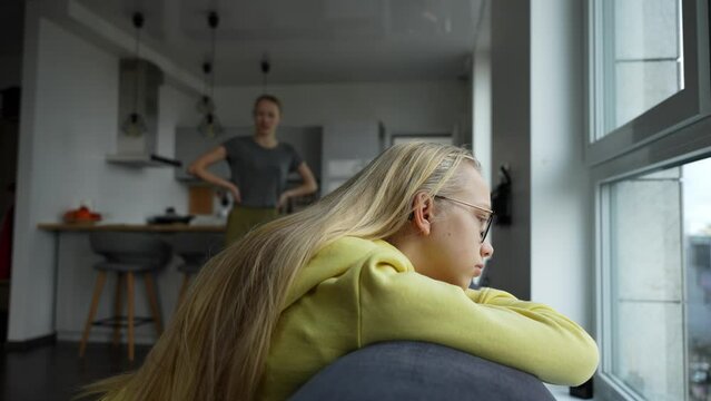 Sad girl portrait. Teen girl listens how her mother is disappointed about daughters behavior. Depressed girl sits on the couch, mother in background.
