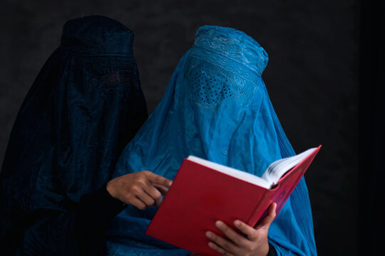 Two Afghan Muslim women with burka tradition cloths in Afghanistan and West Pakistan, studying holy Quran against the dark background