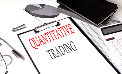 QUANTITATIVE TRADING text on paper clipboard with chart and notebook on the background