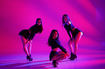 Group of three women posing in a studio in neon pink lighting. Dancers in black shorts and short tops and high heels. Contemporary choreography. Full length. Promo or commercial.