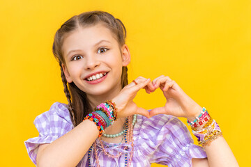 The little girl shows her heart with her fingers and smiles happily. The child put on a lot of beaded bracelets. Beading for children. Yellow isolated background.