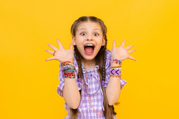 The happy little girl opened her mouth wide with delight and spread her fingers in different directions. The child dressed up in colorful beaded bracelets. Beading for children and teenagers. - obrazy, fototapety, plakaty
