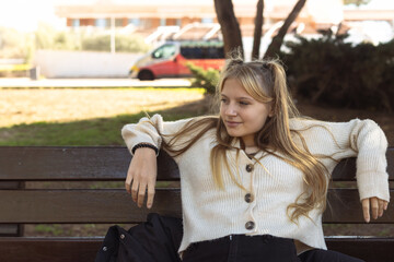 Fototapeta premium a teenage girl of fair appearance with a piercing in the nose dressed in a white jacket, black jeans and red sneakers sits on a bench, close-up on a hipster girl. 