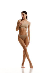 Fototapeta premium Full-length image of slim beautiful brunette woman with relief body standing in beige cotton underwear over white studio background. Concept of beauty, body and skin care, cosmetology, sport, health