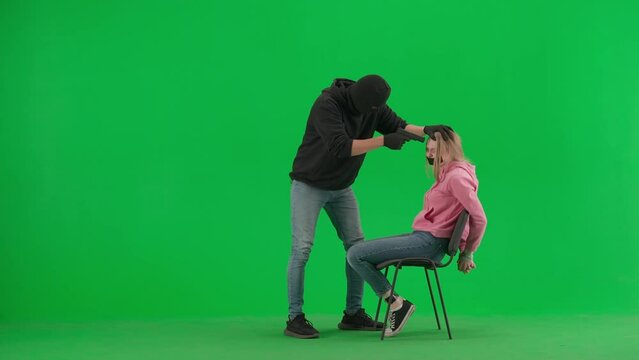 Portrait of thief and victim on chroma key green screen background. Girl sitting on chair tied hands and taped mouth man robber with gun threatens her.