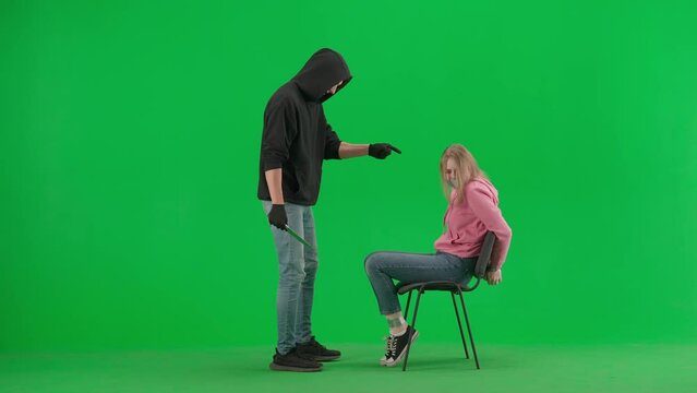Portrait of thief and victim on chroma key green screen background. Girl sitting on chair tied hands and taped mouth man robber with knife threatens her.