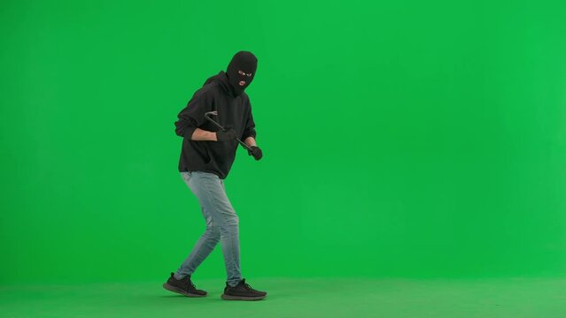 Portrait of thief on chroma key green screen background. Man robber in black balaclava and hoodie walking holding crowbar in hands looks around.