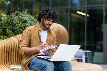 Serious young Muslim man sitting outside on a bench wearing headphones and using a laptop, writing...