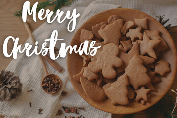 Merry Christmas text on christmas gingerbread cookies in wooden plate on table flat lay with fir branches, pinecone, spices. Happy Holidays! Season's greeting card. Handwritten sign