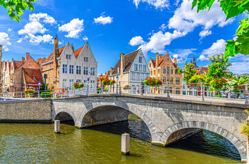 Carmersbrug Carmelite Bridge across Langerei canal, stone arch bridge with flowers on fence, old houses on embankment, Brugge old town district, Bruges city historical center, West Flanders, Belgium - obrazy, fototapety, plakaty