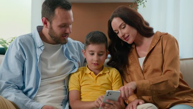 Happy family father mother parents son kid boy child talk communicate looking smartphone screen together on couch home using mobile app for play watch video on Internet browsing social media on phone
