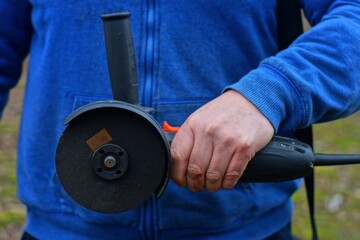 one electric dangerous grinding black industrial grinding machine with a cutting sharp disk in the hand of a worker dressed in a blue raglan on the street during the day