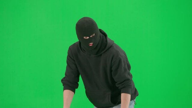 Portrait of thief on chroma key green screen background. Man robber in balaclava and hoodie walking holding gun in hand looks around. Half turn.