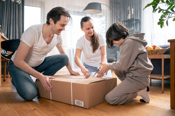 Son, dad and mom, together eagerly unpack a large cardboard box from a parcel with goods that were delivered to their home. Shopping online with home delivery of orders. - obrazy, fototapety, plakaty