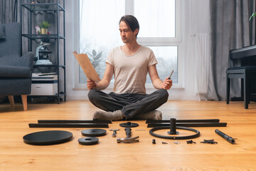 A perfectionist man carefully reads the instruction for assembling furniture, sitting on the floor in the lotus position, among neatly arranged parts and tools. Self-assembly of furniture from scratch - obrazy, fototapety, plakaty