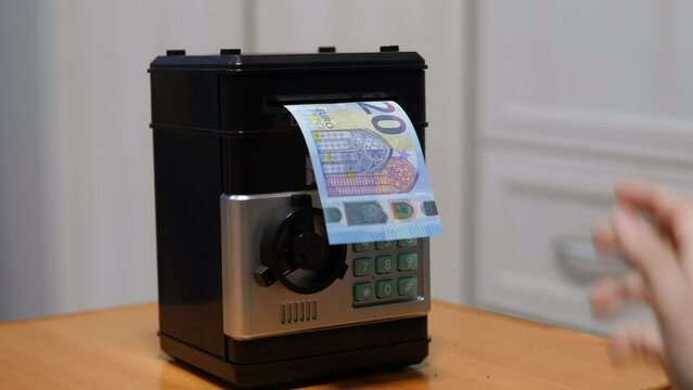 hand of a child lowering euro bills into the bill acceptor of a children's safe at home. European child saves money in a piggy bank safe. schoolchild's pocket money
