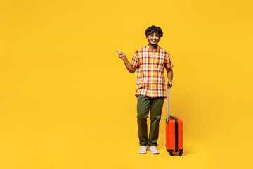 Traveler fun Indian man wear shirt casual clothes hold suitcase point aside isolated on plain yellow background. Tourist travel abroad in free spare time rest getaway. Air flight trip journey concept. - obrazy, fototapety, plakaty