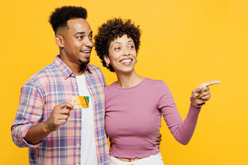 Young fun couple two friend family man woman of African American ethnicity wear purple casual clothes together hold mock up of credit bank card point finger aside isolated on plain yellow background