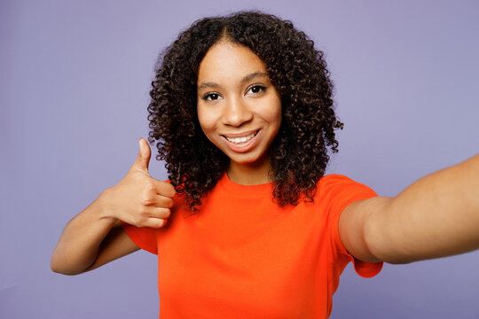 Close up little kid teen girl of African American ethnicity she wear orange t-shirt do selfie shot pov mobile cell phone show thumb up isolated on plain purple background. Childhood lifestyle concept.