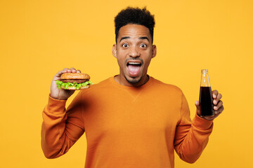 Young amazed man wear orange sweatshirt casual clothes eat fast food burger drink soda pop cola...