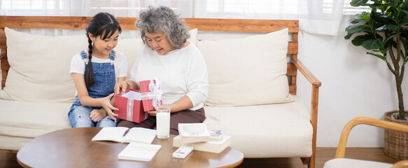 Asian senior woman excited open present box from her granddaughter in the living room. Little girl surprise her grandmother birthday. Love and bonding family. Healthy lifestyle insurance
