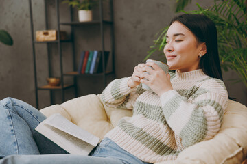 Side view young calm smart fun woman wear casual clothes sits on armchair read book novel drink tea stay at home hotel flat rest relax spend free spare time in grey living room indoor. Lounge concept.