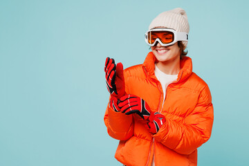Young smiling happy skier woman she wear warm padded windbreaker jacket hat ski goggles mask put on red gloves travel rest spend weekend winter season in mountains isolated on plain blue background.
