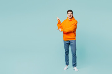 Full body young man wears orange hoody casual clothes point index finger aside on area mock up copy...
