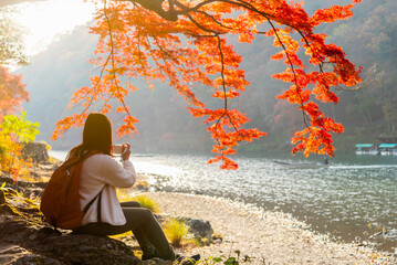 Woman traveler with backpack taking a photo of beautiful nature red maple tree and river in autumn season, Arashiyama Kyoto Japan travel concept.
