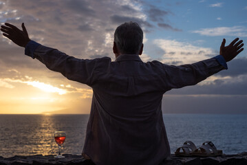 Back view of senior man sitting in front to the sea with outstretched arms looking at sunset,...
