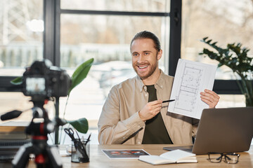 cheerful businessman showing blueprint in front of digital camera during video blog in modern office