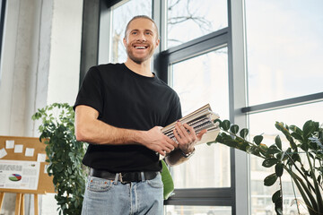 cheerful businessman in black t-shirt holding stack of notebooks and looking away in modern office