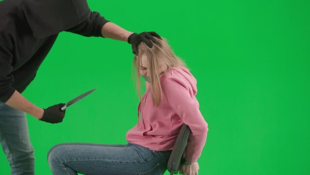 Portrait of thief and victim on chroma key green screen background. Girl sitting on chair tied hands and taped mouth man robber with knife threatens her.