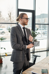 successful businessman in eyeglasses and grey suit standing and looking away in contemporary office