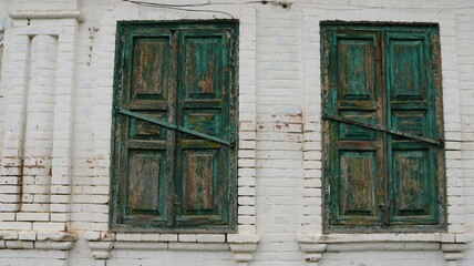 Fototapeta na wymiar two windows of an old whitewashed house bolted with wooden shutters with peeling green paint, fragment of the exterior of an ancient building as a vintage background design, retro texture